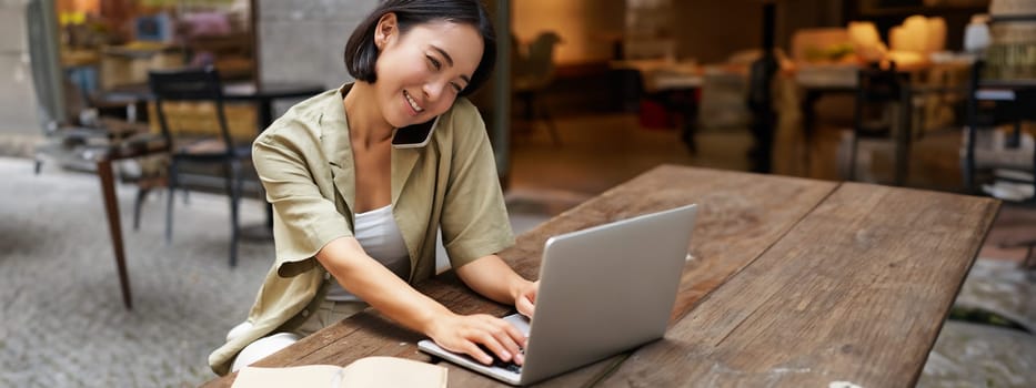 Portrait of young woman talking on mobile phone while typing, using laptop, working remotely from outdoo cafe, doing homework and calling.