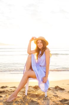 happy beautiful girl in dress and hat sits on chair and looks at sunrise.