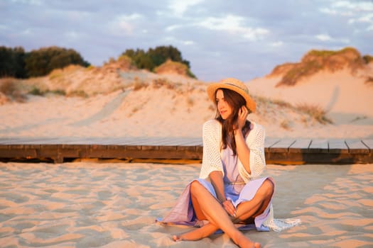 happy beautiful girl in a dress sits on the sand and looks at the sunrise.