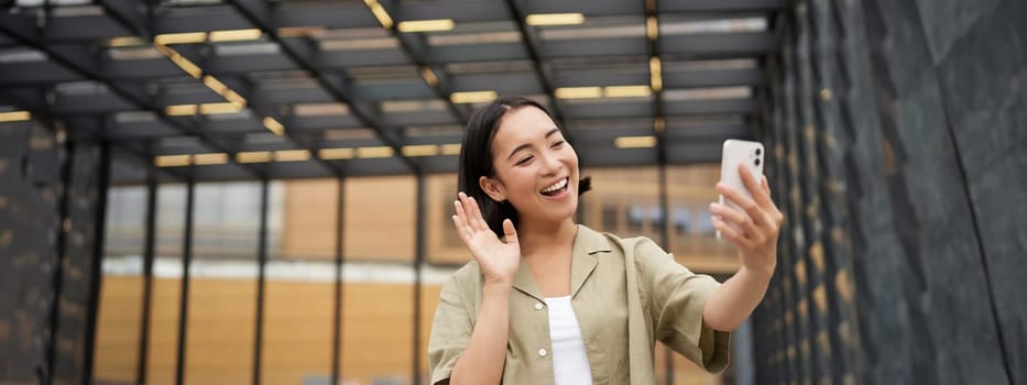 Happy girl says hi to camera on smartphone, video call while stands on street.