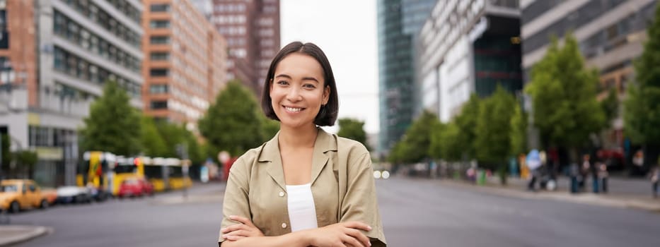 Urban people. Young happy asian girl cross arms on chest, posing on busy city street, smiling with confidence at camera.