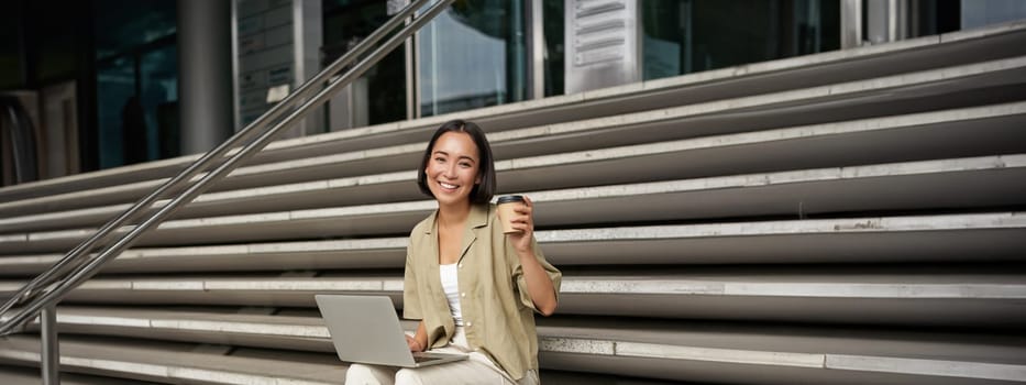 Vertical shot of smiling girl student, asian woman sits on stairs of university campus and drinks coffee, does her homework on laptop.