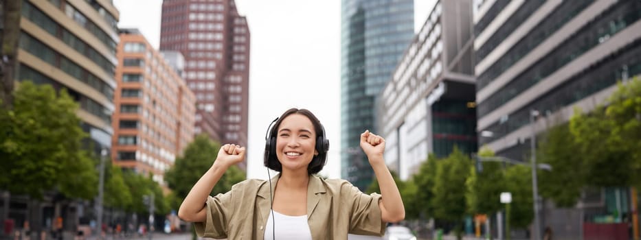 Portrait of smiling asian woman dancing, triumphing, feeling happy while listening music in city, posing on street near skyscrappers.