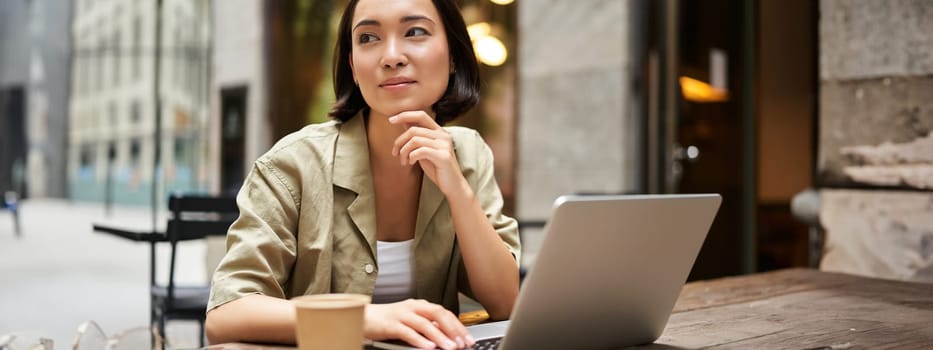 Young woman working in a cafe, using laptop and drinking coffee. Asian girl student with computer studying remotely, sitting on bench near shop.