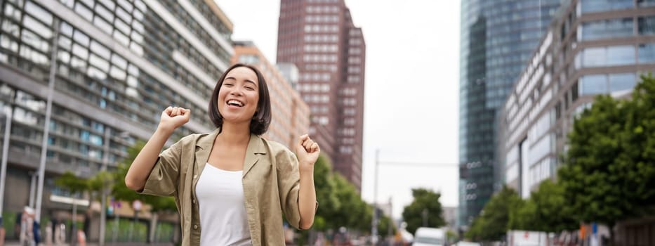 Portrait of happy asian woman, dancing and feeling joy, triumphing, raising hand up in victory gesture, celebrating on streets.