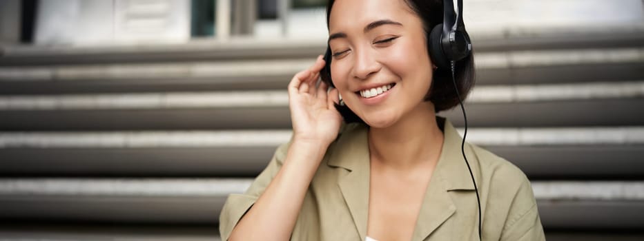 Happy asian girl listens to music in headphones, smiles, sits outdoors on stairs.