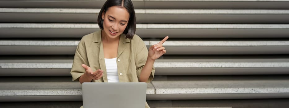 Portrait of young asian girl, student talks at laptop, video chat, speaking during online meeting, sitting outdoors on stairs.