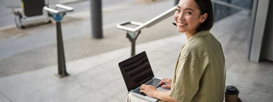 Young smiling woman, asian digital nomad using computer outdoors. Student works on project with laptop, sits on stairs.