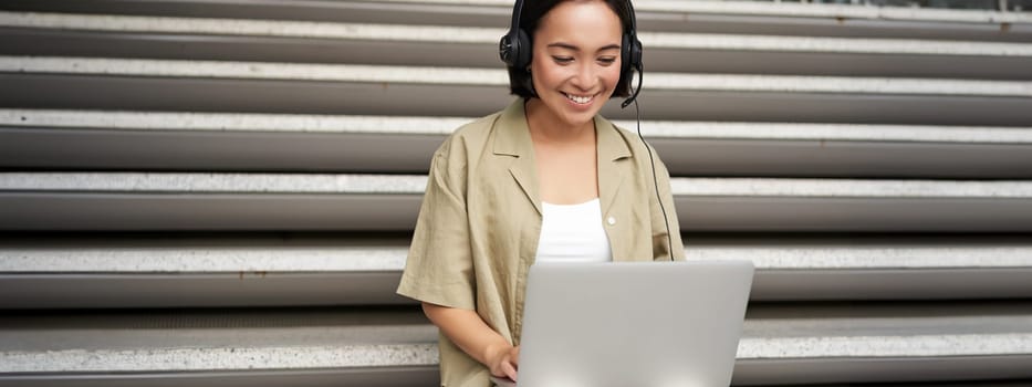 Portrait of asian woman, digital nomad girl using laptop and listens to music outdoors. Young student works on computer and smiles.