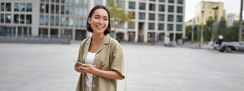 Portrait of asian woman standing on street, city square and holding mobile phone. Girl with smartphone walking outdoors.