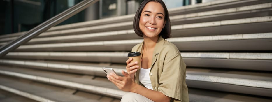 Happy urban girl drinks her takeaway coffee and scrolls feed on smartphone. Asian woman sits on stairs with tea and holding mobile phone.