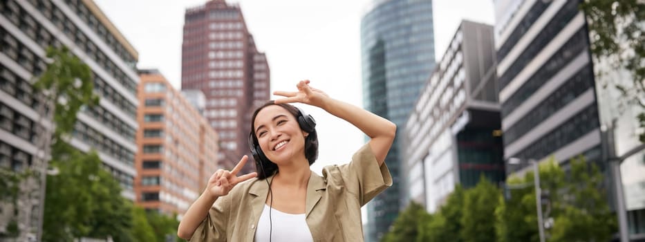 Happy asian woman in headphones, listening music and dancing on street of city centre, smiling with hands up.