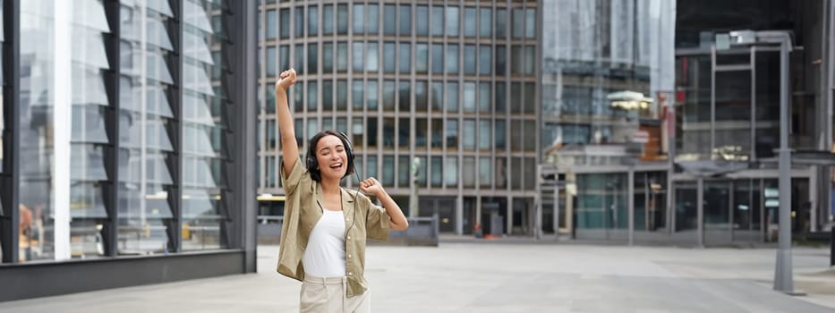 Happy people in city. Upbeat young girl dancing on street in headphones, listening music in headphones.