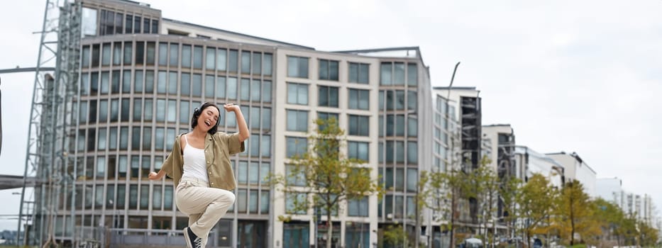 Vertical shot of dancing girl in headphones, listening music on street, enjoying sound in earphones.