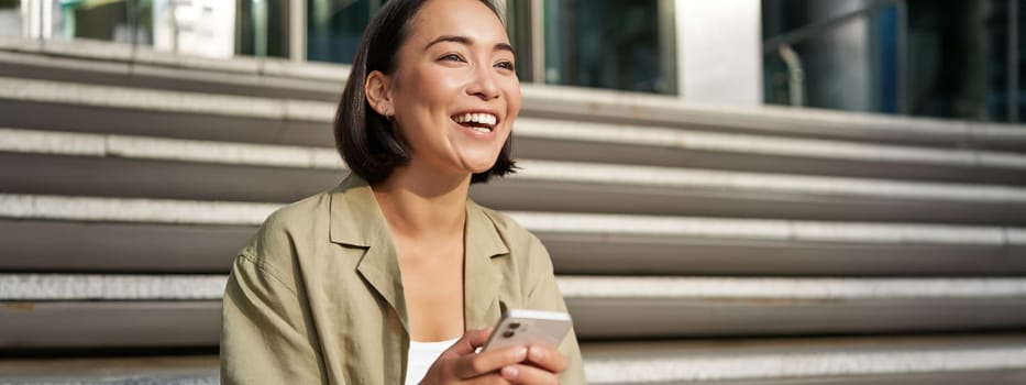 Beautiful asian girl sits on stairs with smartphone. Young korean woman resting outdoors, using mobile phone application.