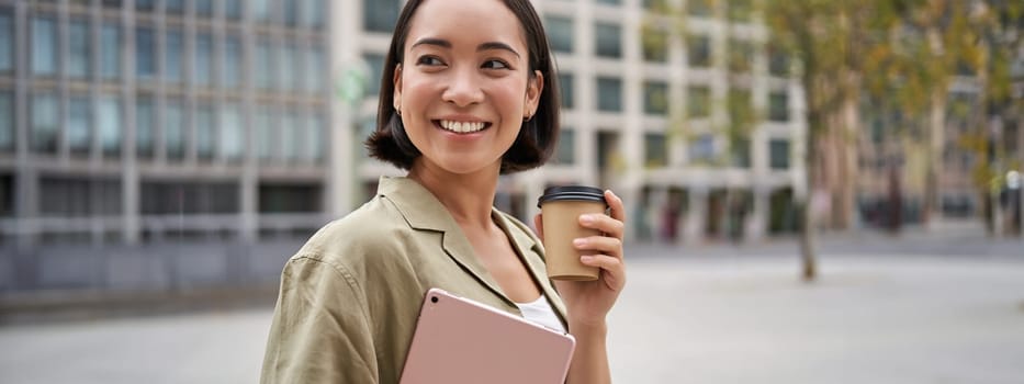 Portrait of asian girl with tablet, drinks coffee on street, walking in city centre.