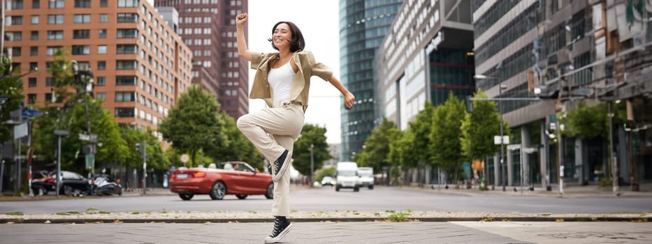 Portrait of asian happy girl jumping and dancing in city centre, posing on streets, express joy and excitement. Copy space