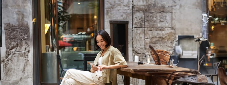 Portrait of stylish young korean woman sits in cafe, holds smartphone, smiles, enjoys coffee outdoors.