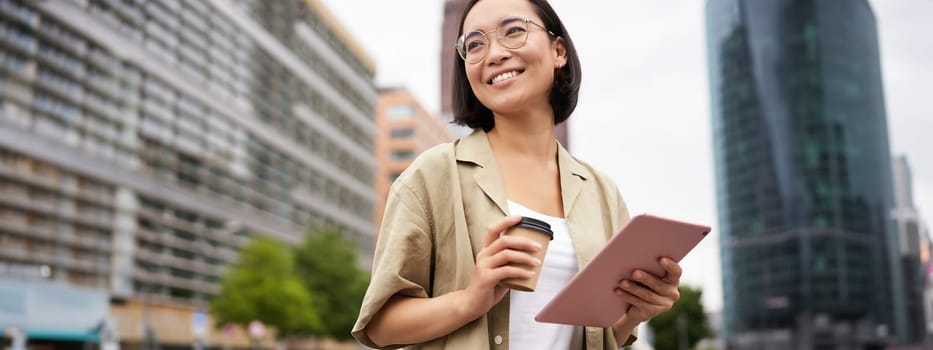 Portrait of happy young woman in glasses, standing on street with cup of coffee and tablet.