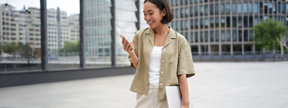 Young asian woman with laptop, walking on street and texting message, smiling while looking at smartphone.