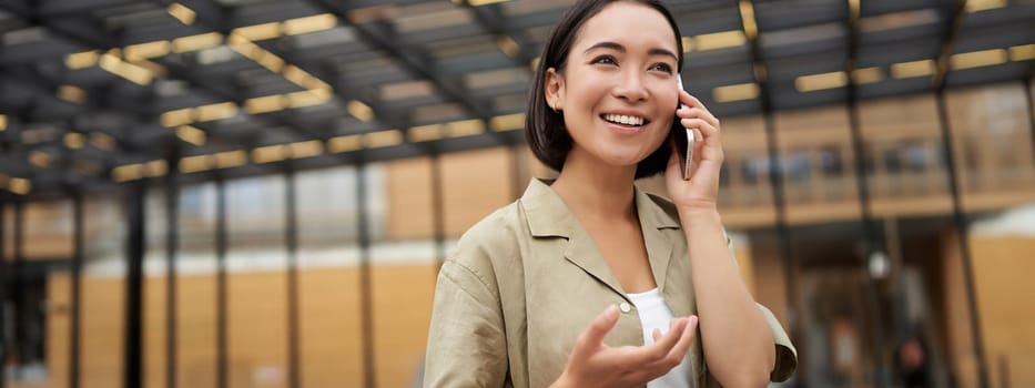 Young asian woman smiles, talks on mobile phone and walks in city centre, poses outdoors.