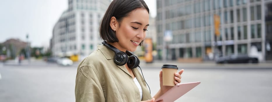 Asian girl walking on street, reading on tablet and drinking coffee outdoors.