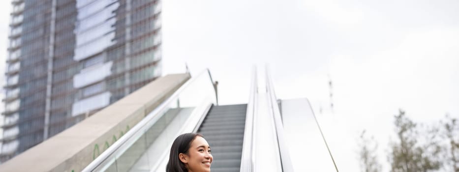 Young asian girl going up on an escalator, holding smartphone, smiling while walking in city. Copy space