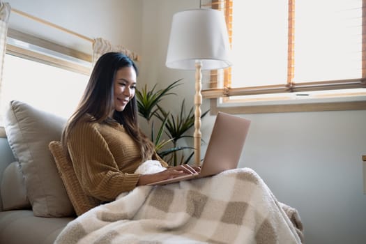 Happy young woman in sweater browsing internet on laptop in a minimalist living room.