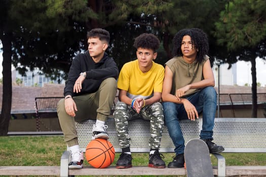 multiracial teen male friends sitting on bench of a park with basketball and skateboard, concept of youth and urban lifestyle