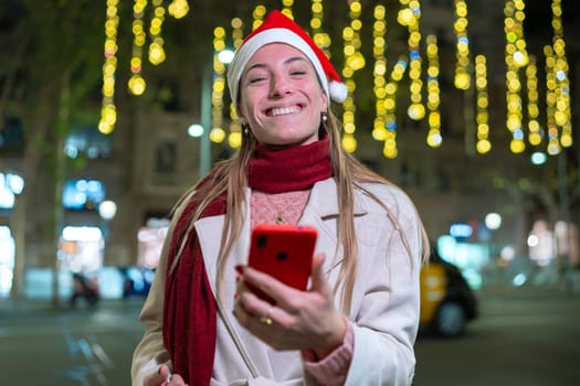 Young woman wearing a Santa hat and red scarf, smiling and holding her smartphone. She stands outdoors at night with festive holiday lights in the background, creating a joyful atmosphere.