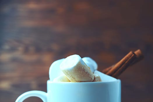 A closeup image of a white mug filled with steaming hot cocoa, garnished with fluffy marshmallows and a cinnamon stick