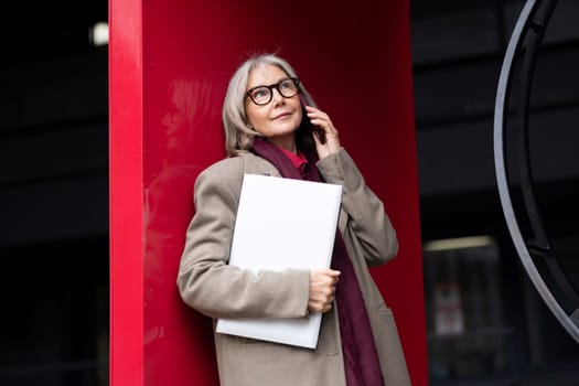 A middle-aged businesswoman stands outdoors, engaging in a phone conversation while holding a laptop close to her. She leans against a large, bright-colored structure.