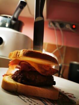 A juicy beef patty topped with crispy bacon is sandwiched between slices of bread, with a knife lodged upright, all set against a home kitchen background.