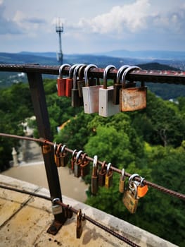 A collection of padlocks in various colors is attached to a metal railing, providing a vibrant contrast against the backdrop of lush trees and distant hills under a clear sky.