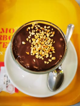 A creamy dessert with a rich chocolate layer and crunchy chopped nuts on top is presented in a glass dish on a white plate. It's a delightful treat for any sweet tooth.