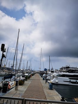 Visitors stroll along the marina walkway, surrounded by a variety of yachts, as clouds loom overhead, creating a calm and peaceful atmosphere in the harbor.