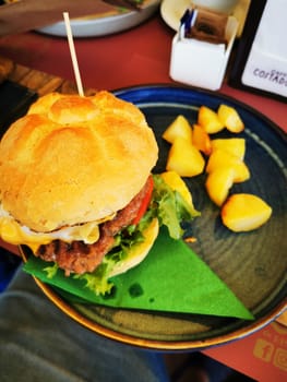 A hearty burger is presented on a plate with crisp lettuce, tomato, and cheese, accompanied by golden potato cubes, all set in a casual dining atmosphere.