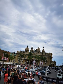Visitors enjoy a vibrant atmosphere around the iconic Monte Carlo Casino, admiring luxury cars while the partly cloudy sky adds to the charm of Monaco.