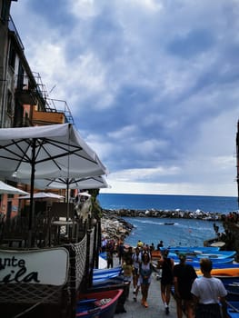 Tourists wander near colorful boats along a coastal walkway, enjoying the view of the ocean and rocky shore while under a partly cloudy sky.