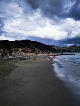 Dark clouds gather over a coastal town, casting shadows on the beach as empty sun loungers line the shore. The calm sea reflects the dramatic sky.