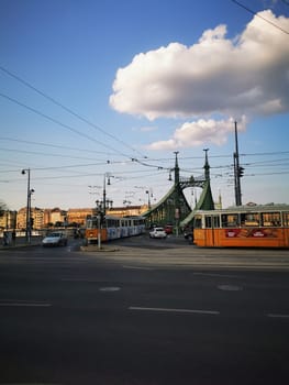 The iconic Liberty Bridge in Budapest stands majestically as trams and cars move through the vibrant city streets under a bright sky with fluffy clouds.