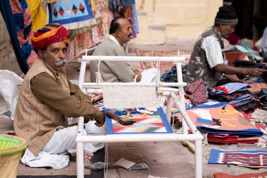 Jodhpur, Rajasthan India - 21st Dec 2023: Indian Rajasthani weaver with colorful turban sitting at a loom creating beautiful clothes in a busy bazaar