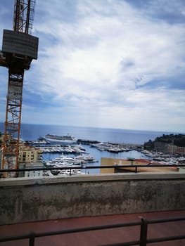 A lively marina filled with numerous yachts is visible beside buildings, while a construction crane stands tall against a backdrop of fluffy clouds and a serene sea.