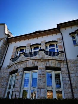 The impressive stone house features multiple windows and a charming balcony. The architecture highlights intricate designs against a bright blue sky, creating a peaceful atmosphere.