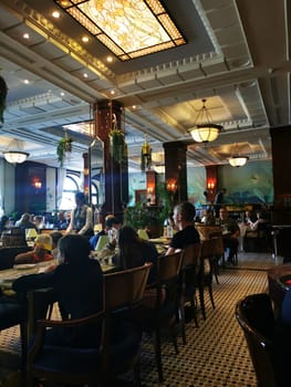 A bustling restaurant filled with patrons engaged in conversation and dining. The elegant interior features ornate lighting and lush greenery, creating a warm ambiance.