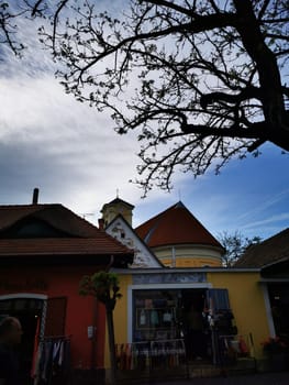 A quaint storefront adorned with colorful architecture sits under a dramatic sky, while tree branches stretch overhead during a serene late afternoon.