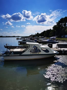 Several boats are moored at a tranquil marina, reflecting on the water's surface. The sky is bright and dotted with fluffy clouds, creating a picturesque setting.