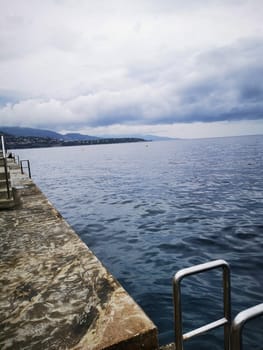A calm body of water stretches out towards a distant shoreline under a blanket of clouds. A paved pier juts out, offering a peaceful spot to enjoy the scenery.