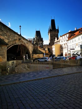 Historic buildings line the cobblestone street leading to the bridge in Prague, showcasing the city's rich architecture under a bright blue sky.