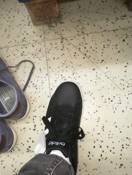 A black sneaker is positioned on a speckled floor, surrounded by other casual shoes. The scene captures a relaxed indoor atmosphere typical of a home or casual space.